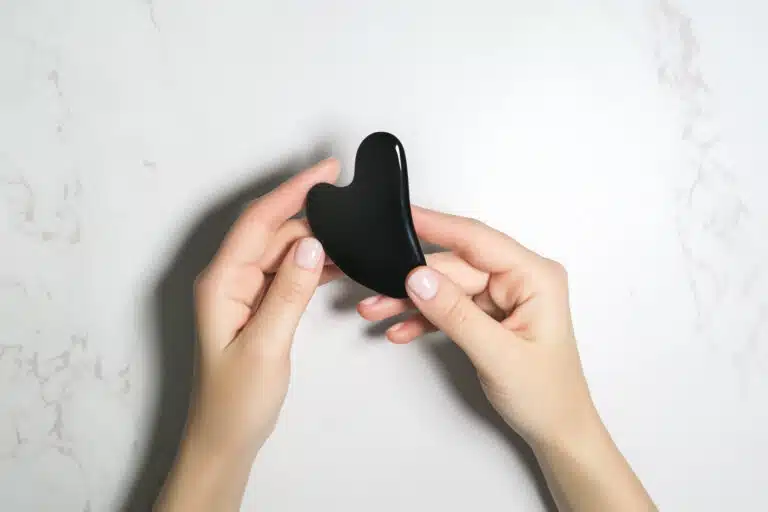 female hands holding a black heart shaped gua sha massager on a marble background, top view